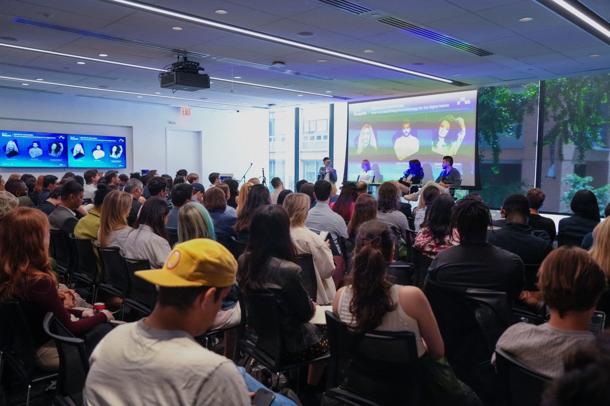 Audience engaged in a panel discussion in a modern seminar room with a projector screen and digital displays.