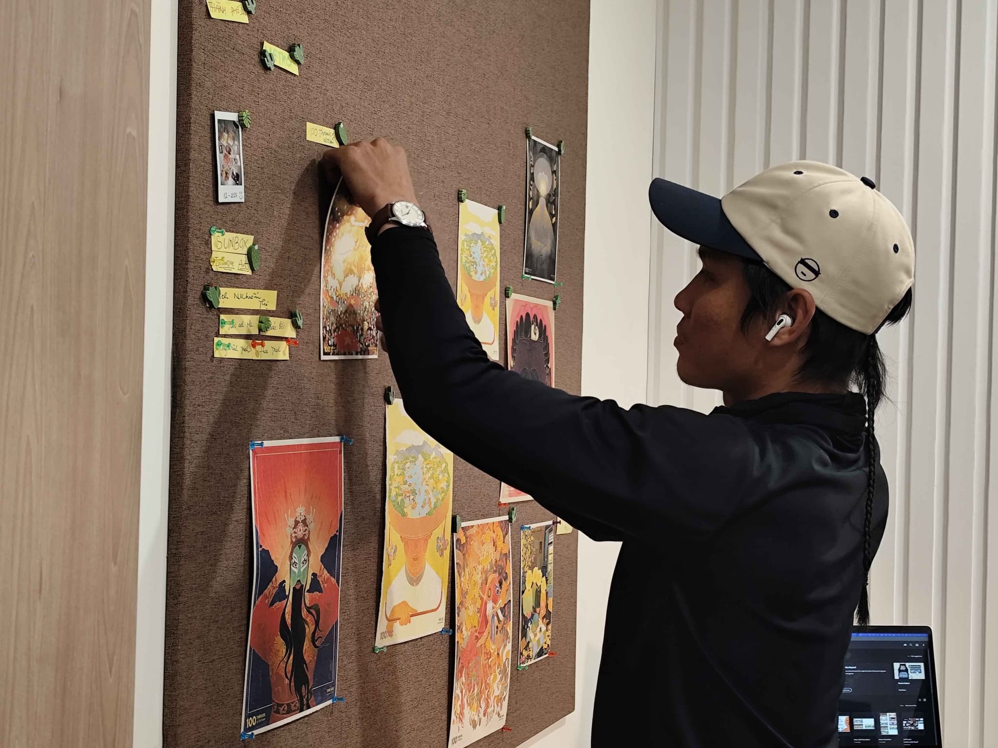Person pinning colorful posters on a corkboard while wearing a cap indoors