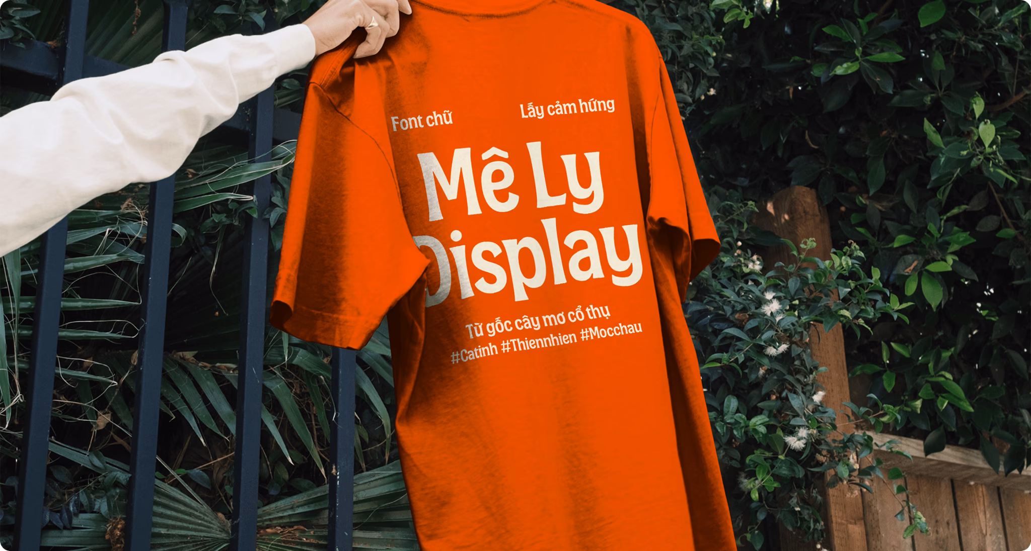 Orange T-shirt displaying "Mê Ly Display" text, showcasing a modern design and vibrant color against a green background.