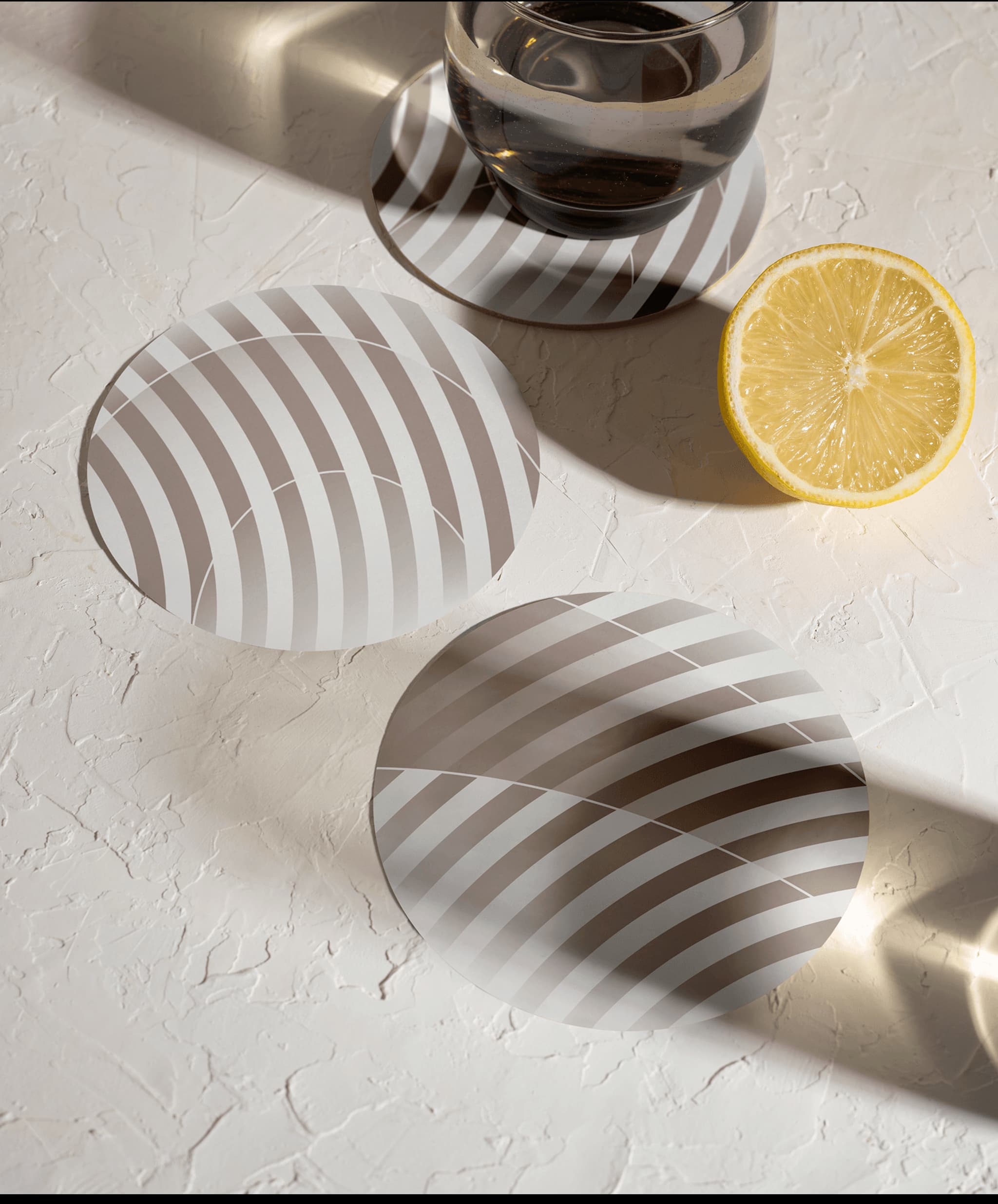 Stylish round coasters with striped design beside a glass of water and a lemon slice on a textured surface.