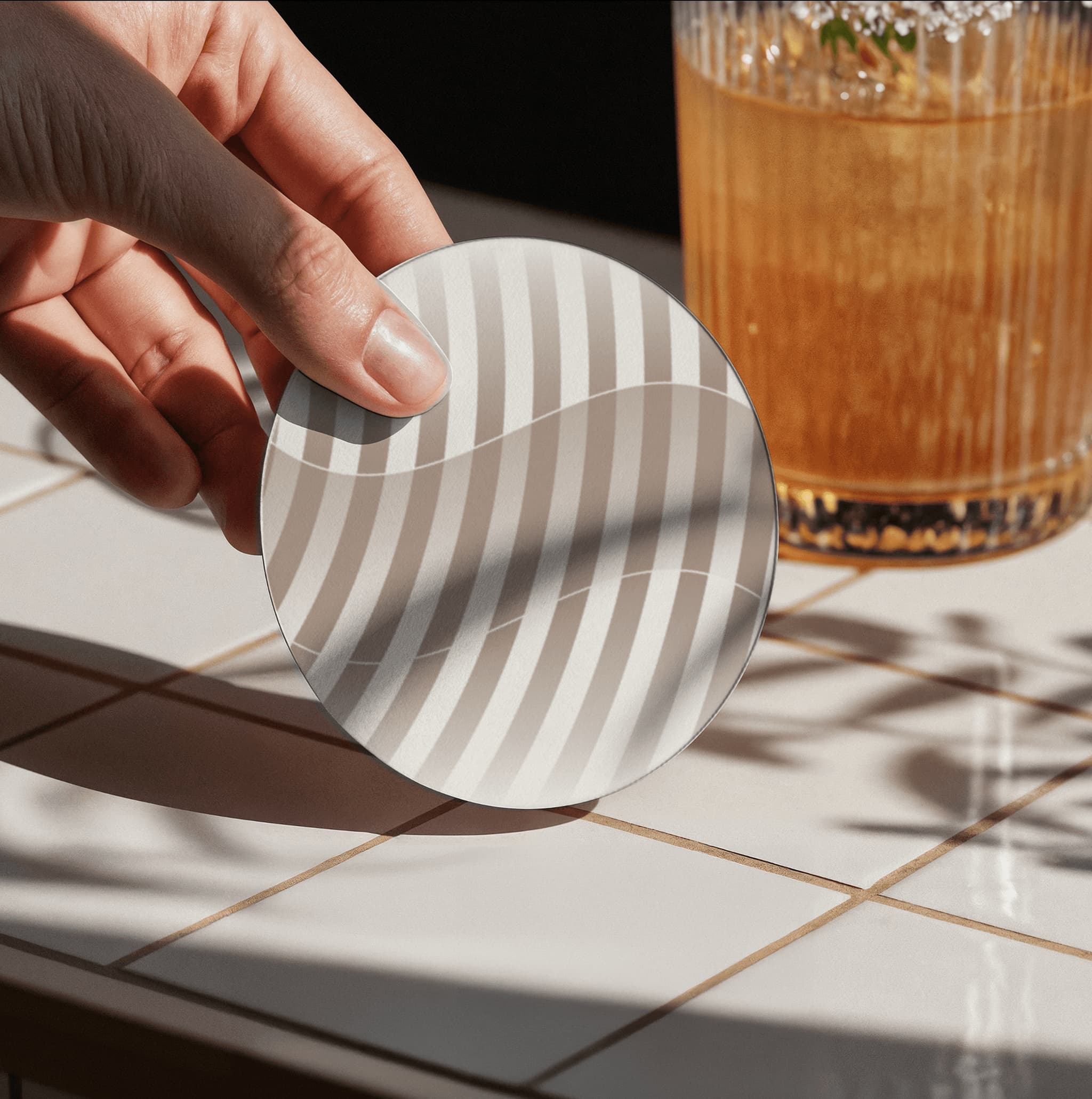 Coaster with wavy pattern held above a tiled surface next to a glass of iced drink.