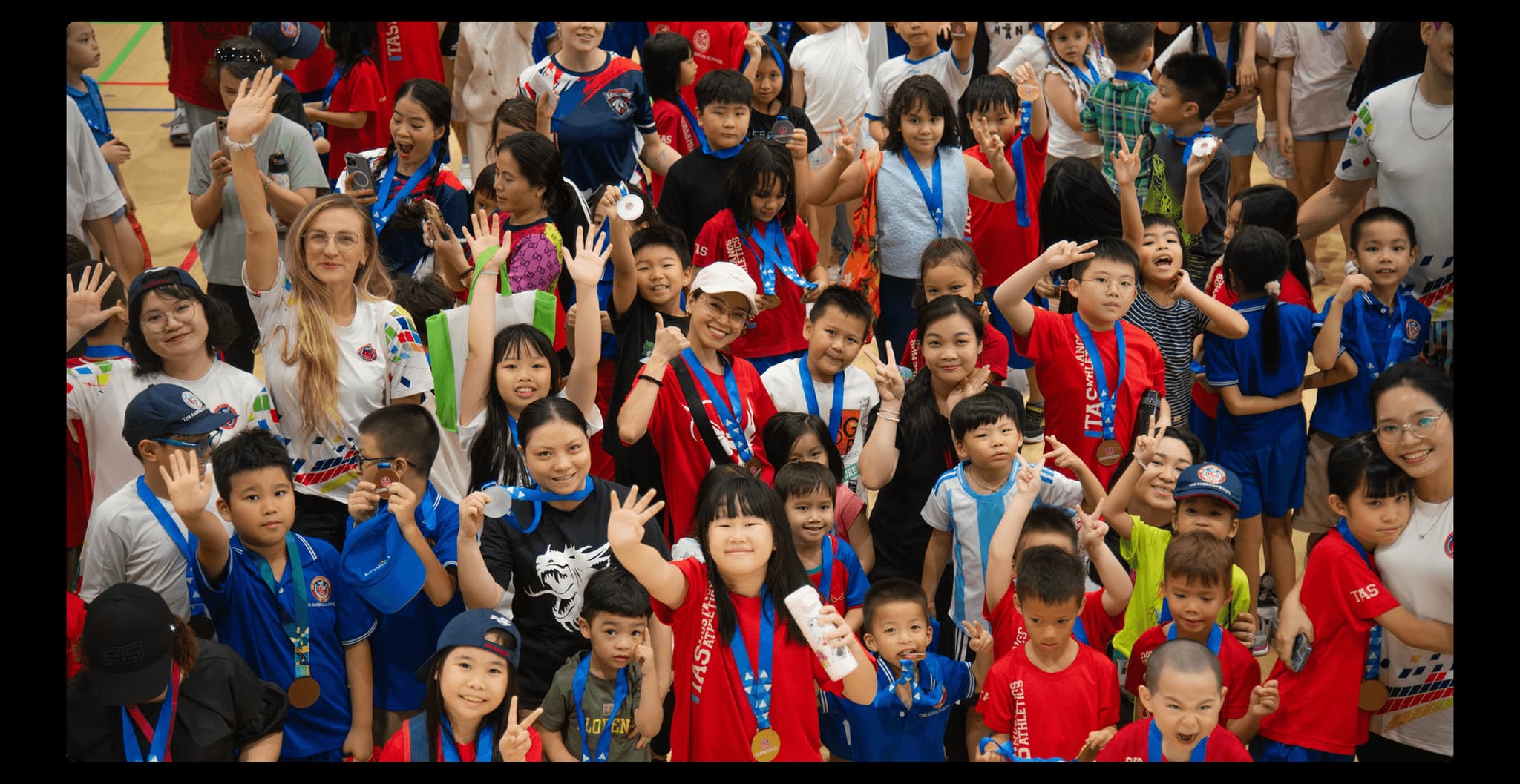 A diverse group of children and adults celebrating with medals, smiling and waving at a sports event.