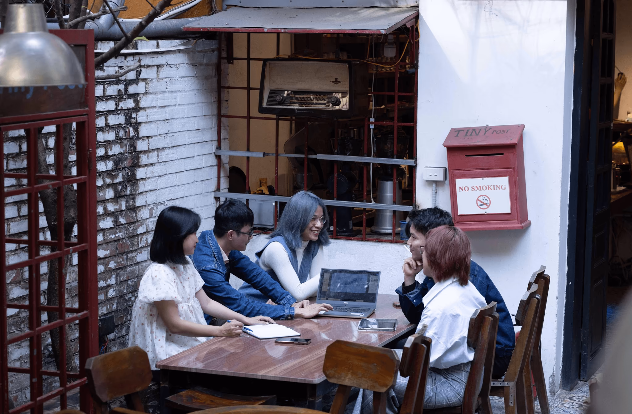 Group of six colleagues collaborating at an outdoor café, engaged in discussion around a laptop.
